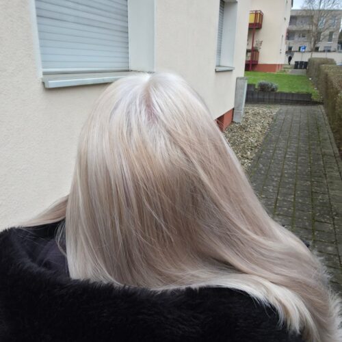 Eine Person mit langen, glatten, platinblonden Haaren steht draußen und trägt eine schwarze Jacke